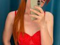 SheritaChieng with red hair, taking a mirror selfie in a red, sparkly dress, holding a beige phone, showing her tits and a slight cleavage.
