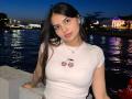 TrishMuhlbauer with long dark hair, white t-shirt showing cherry print, medium tits, standing by water at dusk, looking at camera, blue sky, city lights in background.