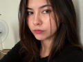 EmeraldCrivaro has straight brown hair, brown eyes, and pink lips. She's wearing a black shirt, looking slightly to the side with a neutral expression. Her face is slightly flushed.