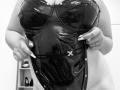 DiLaria porte une combinaison moulante noire en latex brillant, qu'elle tire vers le bas pour exposer ses seins volumineux et brillants, ainsi qu'un petit nœud décoratif. Sa bague est visible à sa main droite..