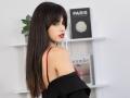JasmineDay with long black hair, red bra straps visible, black off-shoulder top, red lipstick, looking back over shoulder, white shelves with books in background.