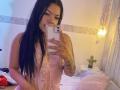AdrianaPaul with long black hair, wearing pink lace lingerie, taking a mirror selfie with her phone. Her tits are visible through the lace, and she has tattoos on her arm.