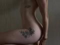 GwynBartell's nude side profile shows a tattooed heart on her ribcage and a rose on her thigh, with a bracelet on her wrist, against a dark background.