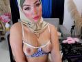 AliceKayleigh with tan skin, green headscarf, pink blush, wearing sheer bra with blue leopard print, exposing her small, pierced tits. She sits in a chair, looking at camera.