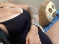 Olieri with large tits in a black dress, tattooed arm, silver watch, and black hair, lying on a blue carpet with a brown teddy bear.
