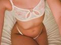 DanikaPlassmann's white lace bra with heart clip, showing her small tits, and white thong, sitting on a bed with her tan, flat stomach and thighs visible.