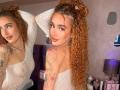 MarissaFex with long curly red hair in a ponytail, white see-through lace bodysuit, tattoos on arms, looking over shoulder, pouty lips, mirror reflection, bedroom setting.