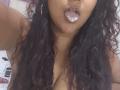 YiyaHot69 with dark curly hair, hoop earrings, and tongue out, showing a bit of white foam on her tongue, and her large, bare, brown, busty chest.