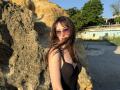 MiraMixi with brown hair, pink sunglasses, and black dress standing against a rocky wall, sunlight highlighting her tits and slender figure, background includes green trees and a blue building.