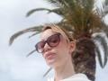 MissFrenchy with short blonde hair, wearing large brown sunglasses, white shirt, and small earrings, standing in front of a palm tree, looking down with a slight smirk.