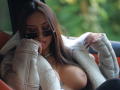 ZenyaNoir with tan skin, brown hair, and big tits, wearing sunglasses, a fluffy beige jacket, and a pearl necklace, resting her chin on her hand, looking down.