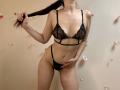 Tisia in black lace lingerie, holding her brown hair, showing her tits and pussy, with her hand covering her crotch, standing against a beige wall with small flowers.