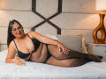 GalileaMoure, Latina with glasses, laying on bed in black bra and fishnet stockings, showing her big tits and fat ass, red lips, long black hair, gold lamp in background.