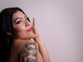 AdrianaPaul with black hair, pink lipstick, and tattoo of a woman with glasses on her shoulder, smiling, hand near face, looking over shoulder, in a studio setting.
