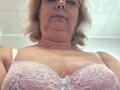 BozhenaNude, middle-aged woman with blonde curly hair, wearing glasses, white lace bra, showing large, lace-covered tits, slightly puffed up, smiling at camera, light skin, gold necklace.