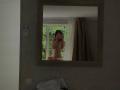 AlexXas, naked, with tits and a pussy, taking a selfie in a mirror, holding a phone, in a dimly lit room, with a curtain and window behind.
