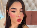 LunaVali met donker haar, een roze blos, opvallende eyeliner, rode lippenstift en een neutrale uitdrukking, zittend op een bed met roze kussens en een gestoffeerd hoofdeinde..