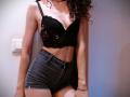 SweetClaraa with curly brown hair, black lace bra, showing small tits, and high-waisted black leather shorts, standing against a white wall, with a light switch to the left.