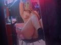 Nude LovelyLith with tits, ass, and pussy, sitting on a chair, arms raised, taking a mirror selfie, wearing black socks, in a dimly lit room.