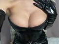 MademoiselleBella's big tits, clad in a shiny black leather corset, with black latex gloves, her hand on her chest, showing her cleavage and a bit of her underboob.