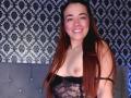 MadisonKoper with long brown hair, smiling, wearing black lace lingerie, exposing small tits with dark nipples, standing in front of a gray tufted headboard.