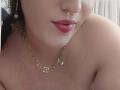 VanessaSquirtt's face, showing shiny skin, red lipstick, and a gold necklace with a small gem and chain links, upper chest visible, black curly hair. Boobs not shown.
