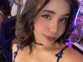 LucieMilner with wavy brown hair, wearing a blue floral top and black choker, has a cute face, pink blush, and a slight smile, with small tits visible.