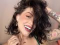 HannaBorbon has messy black hair, a tattooed arm, and is smiling with dark lipstick, showing white teeth. She's wearing gold earrings and a green top.