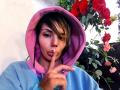 MissLover portant un sweat à capuche rose et bleu, un doigt sur les lèvres pour faire signe de silence, avec des cheveux bruns, des yeux roses et un regard séduisant, entourée de roses rouges et de feuilles vertes..