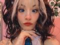 LaurenEvie69 with blue eyes, white and black wavy hair, pink nose, and orange lips, hand on neck, visible tattoo on wrist, wearing blue plaid tie, pink room.
