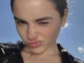 PeacHub69's face with a small, noticeable scar above their right eyebrow, slightly puckered lips, and dark hair tied back. Wearing a black leather jacket, blue sky in background.
