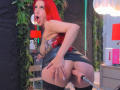 AlissaBrown with bright red hair, fishnet top, and black leather skirt, bent over, showing her ass with a dildo inserted, looking surprised with mouth open.