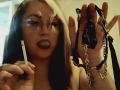 BeccaVanBoa with glasses, black lipstick, holding a cigarette in one hand and black leather chains with metal rings in the other, her tongue slightly out.