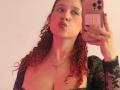 ValeryScot with red curly hair, puffy lips, holding a pink phone, wearing a black lace top, exposing her tits, and a necklace, against a white wall.
