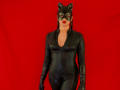 AdalinaVera in a black, shiny, tight-fitting latex catsuit with cat ears mask, red lipstick, and black gloves, standing against a bright red background. She's got a cute ass.