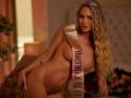 Nude RosalieFleur with long blonde wavy hair, pink tiara, and gold sash over her tits, leaning forward, showing her ass and tattoos, in a warm, dimly lit room.