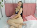AbbyHillsI with green and black hair, smiling, wearing navy blue lace bra and thong, sitting on pink couch with pink pillows, legs crossed, showing her ass and pussy.