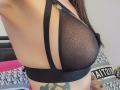 FrenchShanna's tits in a sheer black bra with dotted mesh, showing a small silver ring. Her tattooed side and long brown hair are visible.