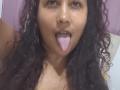 YiyaHot69 with curly dark hair, brown skin, and small hoop earrings, sticking out a pink tongue. Her nose is slightly upturned. She's wearing a sleeveless top.