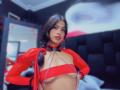 MelisaDaltonn with red mesh top, black choker with heart, chains across tits, dark hair, pink lipstick, standing in bedroom, black headboard, mirror, and red string lights in background.