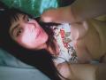 AnyMadison with long black hair, wearing a floral top, exposing her tits, lying down, pouty expression, green bedsheet in background, soft light highlighting her face and chest.