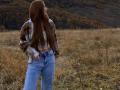 JaneKrover with long brown hair, plaid jacket, and blue jeans stands in a grassy field, hands in pockets, looking to the side, her ass slightly visible.
