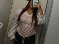 AntoniaBrunni with long red hair, smiling, wearing a pink floral top with white puffed sleeves and black pants, taking a mirror selfie in a dimly lit bathroom.