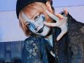 ViolettClay with painted white face, black lipstick, and blue makeup under eyes. She's making a peace sign, wearing a black lace top and beret, with pinkish-brown hair.