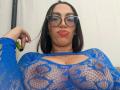 CamilleJuicyHot wearing blue lace top, showing big boobs through mesh, with glasses and pink lipstick, holding green pen in right hand, brown hair, white background.