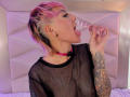 DannyRouds with pink-dyed hair, black mesh top, pink choker, and tattooed arm, licking a transparent glass dildo. Bedhead in background.