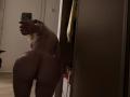 Nude MillieHartwell with a large ass, standing in a dim room, taking a selfie with her phone, showing her back and buttocks, blonde hair, in front of a mirror.