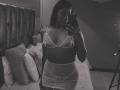 Amberv with dark hair, wearing sheer lingerie with small tits and a visible pussy, takes a mirror selfie in a dimly lit room. She holds a phone in her right hand.