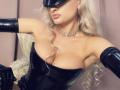 MademoiselleBella with platinum blonde hair, black latex corset, and gloves, wearing a blindfold, choker, and earrings, showing her tits and pink lips. Her ass is not visible.