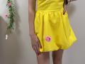 MiraMixi wearing a yellow dress with a pink flower on the right side, red nails, hand on her hip, white wall background, green vine with pink flowers on left.
